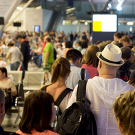 Crowd of travellers queuing inside a busy airport terminal, seen from behind, with a man in a white hat and light hooded top in the foreground.
