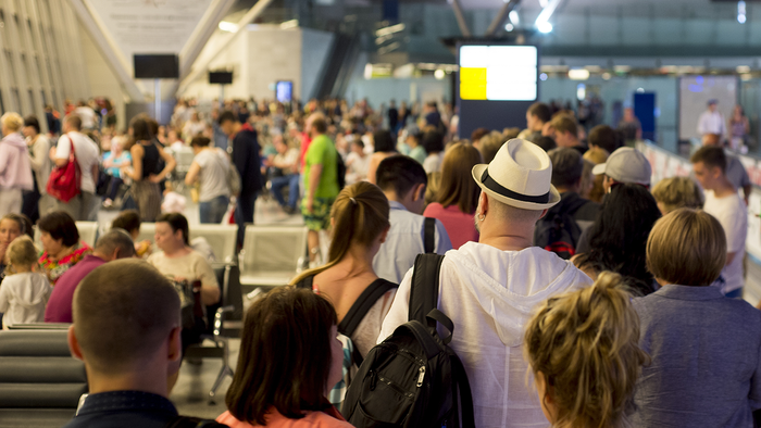 Crowd of travellers queuing inside a busy airport terminal, seen from behind, with a man in a white hat and light hooded top in the foreground.