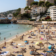 Crowded beach in Mallorca with sunloungers and parasols along the shoreline, swimmers in the water and apartment buildings on the hillside behind.