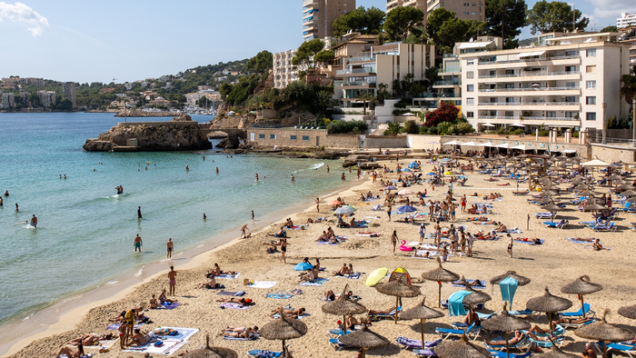 Crowded beach in Mallorca with sunloungers and parasols along the shoreline, swimmers in the water and apartment buildings on the hillside behind.