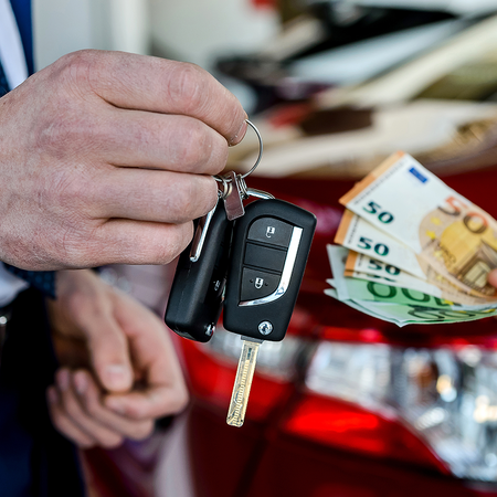 A man in a blue suit holds out a car key while another person counts euro banknotes beside a red car.