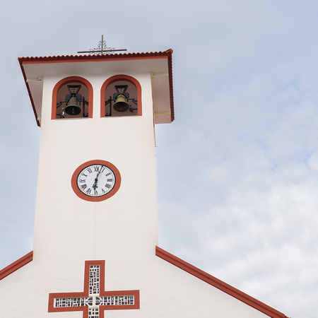 A white bell tower with a clock and two bronze bells under a red-tiled roof, against a partly cloudy sky.