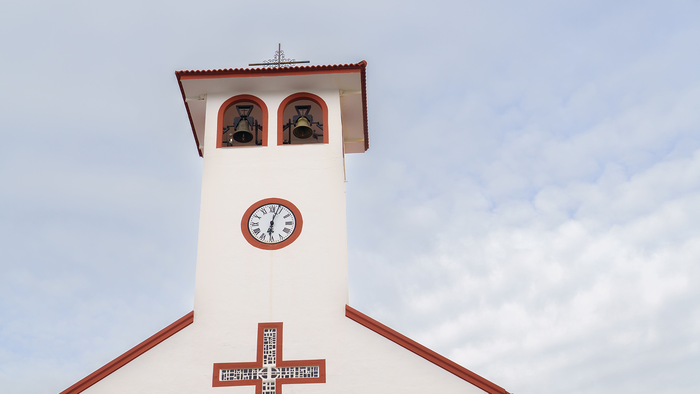 A white bell tower with a clock and two bronze bells under a red-tiled roof, against a partly cloudy sky.