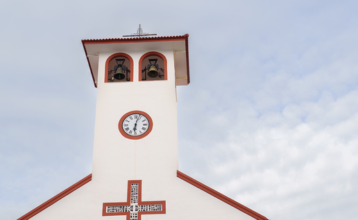 A white bell tower with a clock and two bronze bells under a red-tiled roof, against a partly cloudy sky.