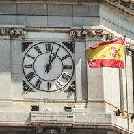 A stone clock tower with Roman numerals and a Spanish flag flying beside it against a blue sky.