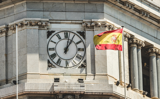 A stone clock tower with Roman numerals and a Spanish flag flying beside it against a blue sky.