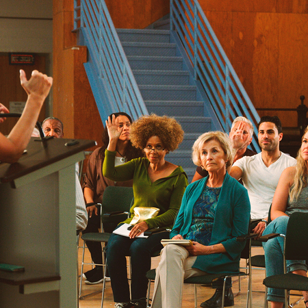 A woman speaks from a podium to a seated audience, several of whom have their hands raised with questions.