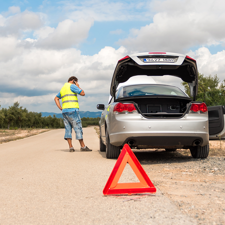 A driver in a high-visibility vest talks on the phone beside a silver convertible with its boot open, with a red warning triangle on a rural road lined with olive trees.