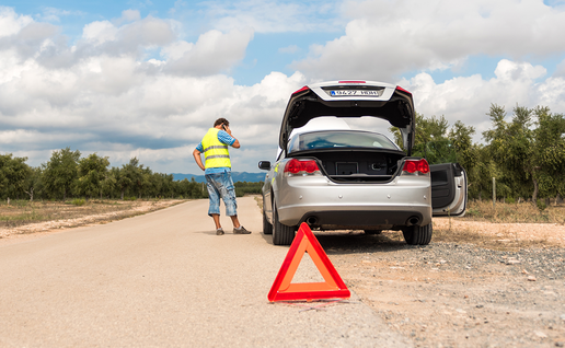 A driver in a high-visibility vest talks on the phone beside a silver convertible with its boot open, with a red warning triangle on a rural road lined with olive trees.