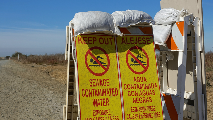 A yellow bilingual sign warning of sewage-contaminated water and prohibiting swimming, mounted on a barrier at a beach access point.