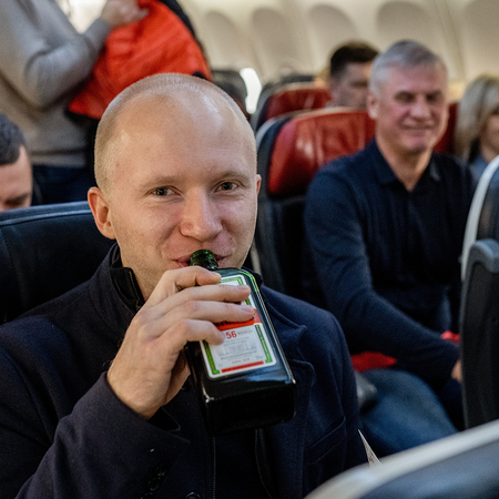 A passenger drinks from a small bottle of spirits while seated among other passengers on a plane.