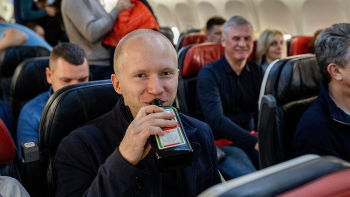 A passenger drinks from a small bottle of spirits while seated among other passengers on a plane.