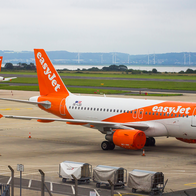 Two orange and white easyJet Airbus A320 aircraft parked on an airport apron, with wind turbines and hills visible in the distance.