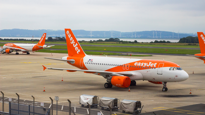 Two orange and white easyJet Airbus A320 aircraft parked on an airport apron, with wind turbines and hills visible in the distance.