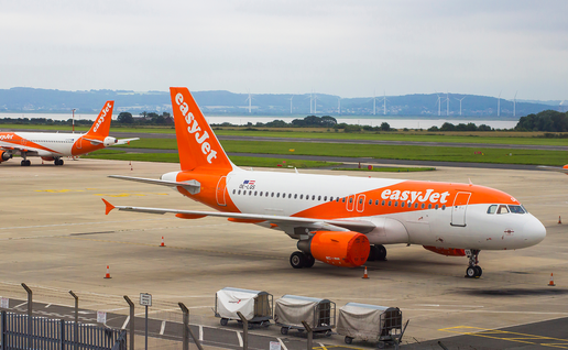 Two orange and white easyJet Airbus A320 aircraft parked on an airport apron, with wind turbines and hills visible in the distance.
