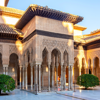 An ornate Alhambra courtyard in Granada, with carved Moorish arches, marble columns and two small trees lit by late afternoon sun.