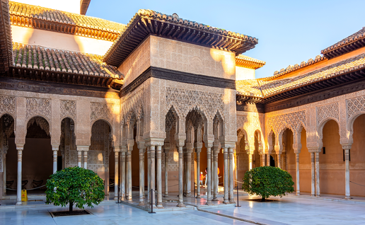 An ornate Alhambra courtyard in Granada, with carved Moorish arches, marble columns and two small trees lit by late afternoon sun.