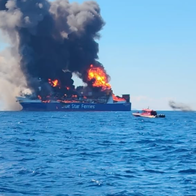 A large car ferry marked "Blue Star Ferries" engulfed in flames and thick black smoke at sea, with two small rescue boats standing off nearby.