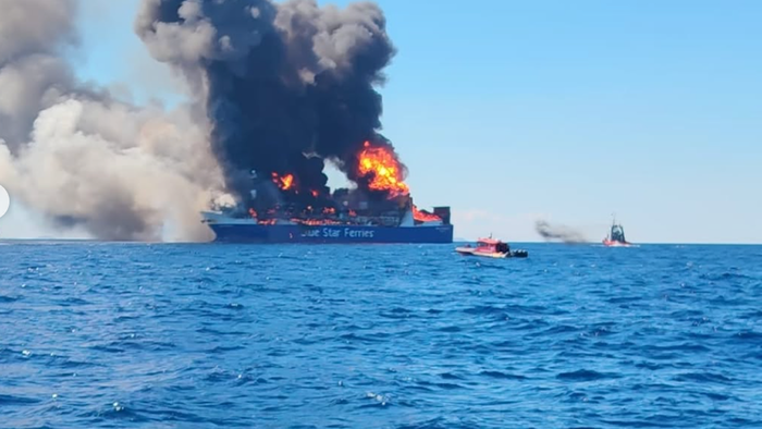 A large car ferry marked "Blue Star Ferries" engulfed in flames and thick black smoke at sea, with two small rescue boats standing off nearby.