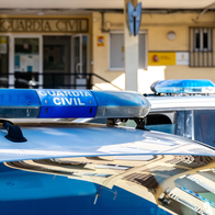 Close-up of a Guardia Civil patrol car's blue light bar, with a second marked vehicle and the entrance to a Guardia Civil station visible behind it.