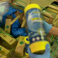 A gloved hand holds a yellow-capped canister with a blurred label in front of cardboard boxes, wooden pallets and blue plastic sheeting, with a Guardia Civil logo in the top left corner.