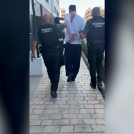 Two Guardia Civil officers escort a handcuffed man down a paved walkway between apartment buildings.