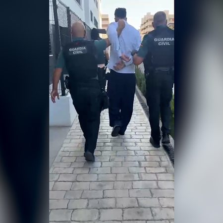 Two Guardia Civil officers escort a handcuffed man down a paved walkway between apartment buildings.