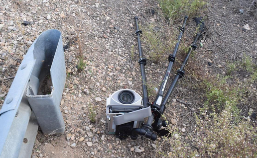A damaged mobile speed camera and its tripod lying on gravel beside a road guardrail.
