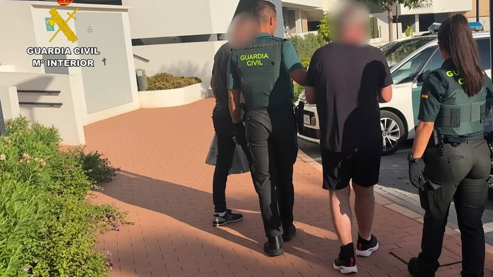Guardia Civil officers escorting two people with blurred faces outside a residential building.