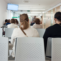 Patients seated in rows of waiting room chairs in a hospital corridor, seen from behind.