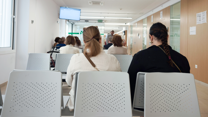 Patients seated in rows of waiting room chairs in a hospital corridor, seen from behind.