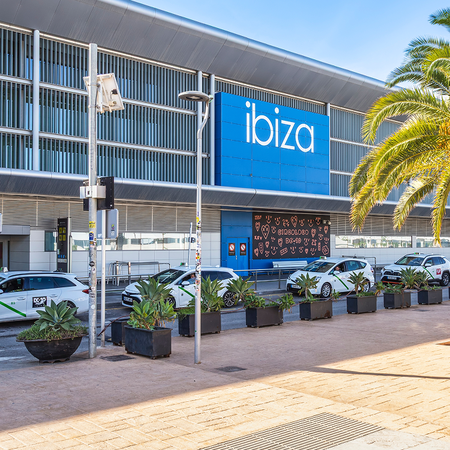 Exterior of Ibiza Airport's arrivals terminal, with the blue "ibiza" sign, palm trees and taxis parked outside on a sunny day.