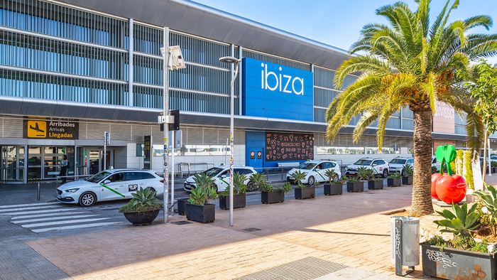 Exterior of Ibiza Airport's arrivals terminal, with the blue "ibiza" sign, palm trees and taxis parked outside on a sunny day.