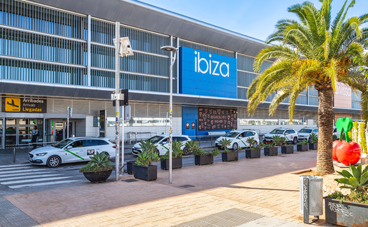 Exterior of Ibiza Airport's arrivals terminal, with the blue "ibiza" sign, palm trees and taxis parked outside on a sunny day.