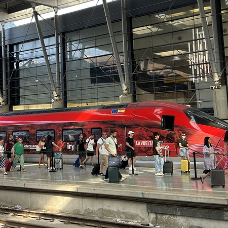 Travellers with suitcases walk along a platform beside a red high-speed train at a Spanish railway station.