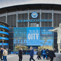 Fans walking past Manchester City's Etihad Stadium, with the club's crest and a player statue outside the ground.
