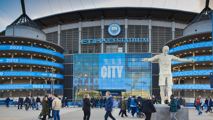 Fans walking past Manchester City's Etihad Stadium, with the club's crest and a player statue outside the ground.