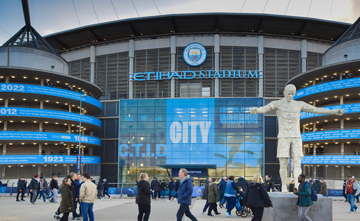 Fans walking past Manchester City's Etihad Stadium, with the club's crest and a player statue outside the ground.