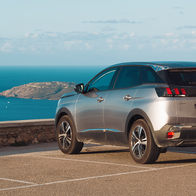 A car parked at a clifftop viewpoint overlooking a coastal bay in Menorca, with a marina and village visible below.