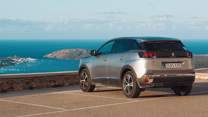 A car parked at a clifftop viewpoint overlooking a coastal bay in Menorca, with a marina and village visible below.