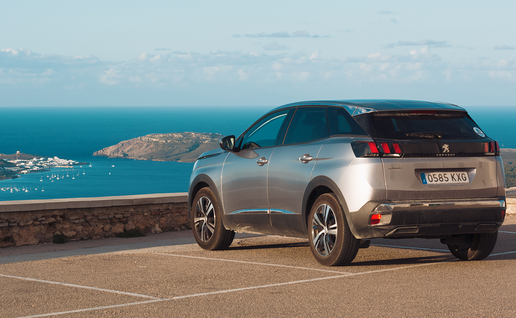 A car parked at a clifftop viewpoint overlooking a coastal bay in Menorca, with a marina and village visible below.