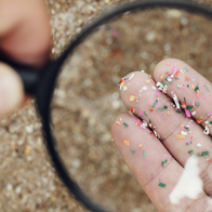 A magnifying glass held over sand reveals brightly coloured microplastic fragments scattered across a person's fingers