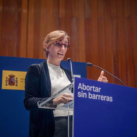 Mónica García speaks at a blue podium reading "Abortar sin barreras", in front of a wood-panelled wall and a blue backdrop with the Spanish government crest.
