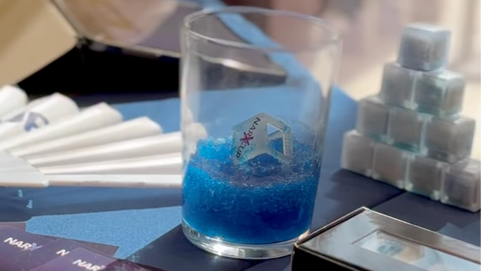 A NarXcup-branded ice cube stirrer sitting in a glass of blue, gelled drink, next to a stack of unused NarXcup ice cubes and branded promotional cards on a table.