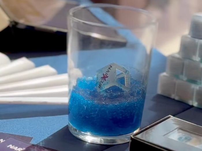 A NarXcup-branded ice cube stirrer sitting in a glass of blue, gelled drink, next to a stack of unused NarXcup ice cubes and branded promotional cards on a table.