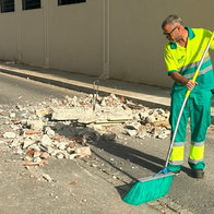 Man in high-visibility green uniform sweeping rubble on a street with a long-handled broom.
