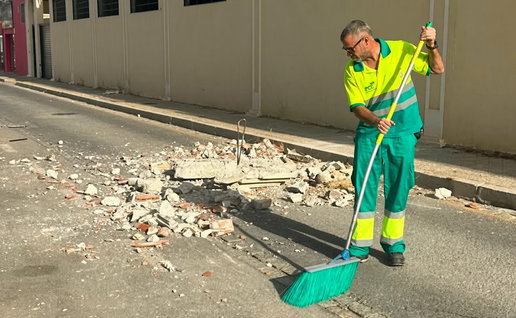 Man in high-visibility green uniform sweeping rubble on a street with a long-handled broom.