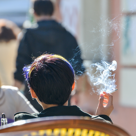 Person holding a lit cigarette while sat on a bar or restaurant terrace, with other people sat on other tables near them.