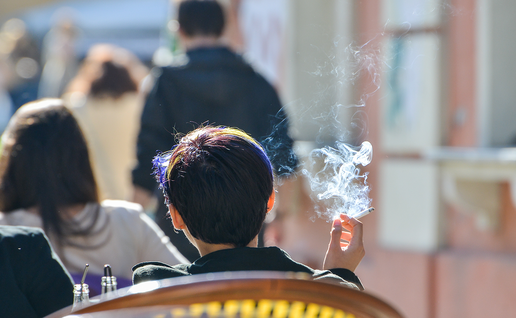 Person holding a lit cigarette while sat on a bar or restaurant terrace, with other people sat on other tables near them.