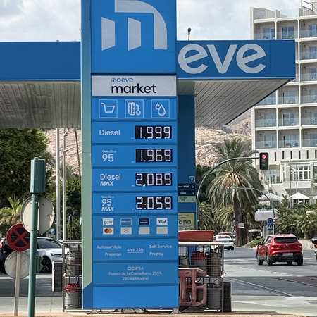 An electronic fuel price sign at a Moeve service station in Almería showing diesel and petrol prices per litre, with traffic and a hotel in the background.